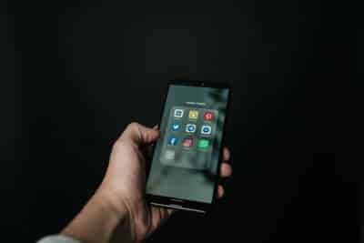 Close-up of a hand holding a smartphone displaying various social media app icons on a dark background.