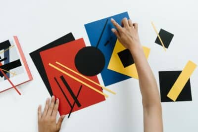 Hands arranging colorful geometric paper shapes in an abstract modern art style.